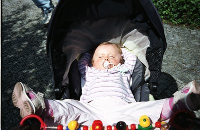 A baby lies asleep in a stroller, pacifier in mouth, wearing a pink and white striped outfit with socks and shoes. The stroll...