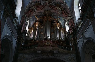 Ornate, gilded pipe organ dominates a baroque church interior. Elaborate carvings & painted frescoes featuring cherubic figur...