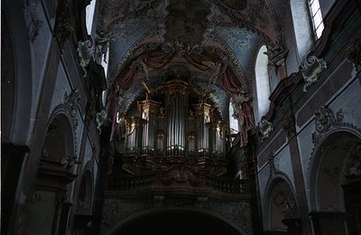 Ornate Baroque organ dominates a church interior. Elaborate carvings, dark wood, and painted ceiling with cherubic figures cr...