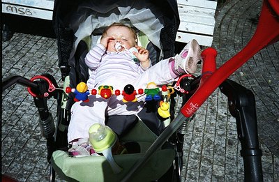 Infant asleep in a green stroller, pacifier visible. Red stroller frame, baby toys dangling above. Likely a candid, casual sn...