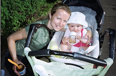 A young woman smiles while feeding a baby in a stroller. The baby wears a bonnet and bib, gazing towards the camera. The scen...