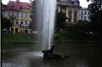 A tiered fountain with a sculpted figure spouts water in a central pond. Behind, ornate, multi-story buildings with peaked ro...