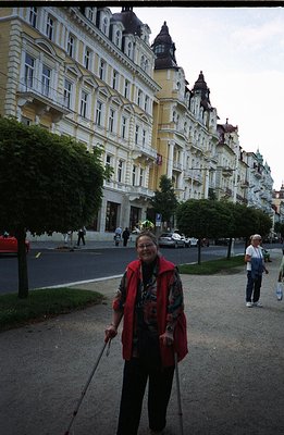 A woman with walking sticks stands on a paved walkway facing a grand, multi-story building with ornate balconies and arched w...