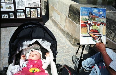 A baby sleeps peacefully in a stroller, positioned near a seated artist working on a landscape painting depicting a building ...