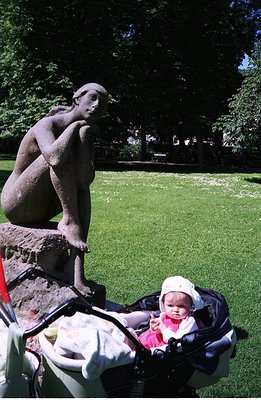 A bronze sculpture of a woman seated on a rocky base is framed by lush greenery. A baby sits in a stroller in the foreground,...