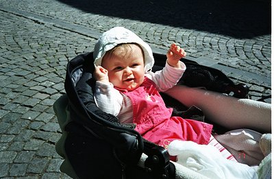 A smiling infant sits in a stroller, dressed in a pink dress and white bonnet. The background shows a stone-paved surface. Li...