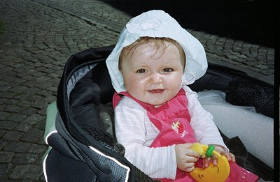 Infant in a stroller, wearing a white bonnet and pink dress. Holding a rubber duck toy. Likely a family snapshot, potentially...