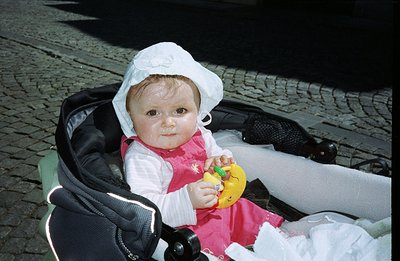 Infant in a stroller, wearing a white bonnet and pink dress. Holds a yellow duck toy. Likely a candid portrait, possibly a fa...
