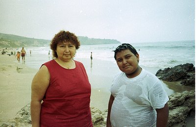 A woman and boy stand on a sandy beach, likely a family vacation snapshot. The woman wears a red sleeveless dress; the boy, a...
