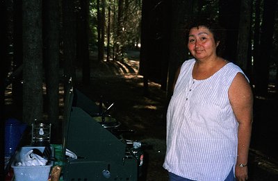 A smiling woman stands outdoors beside a camping stove and equipment, possibly for cooking. Forested background with filtered...