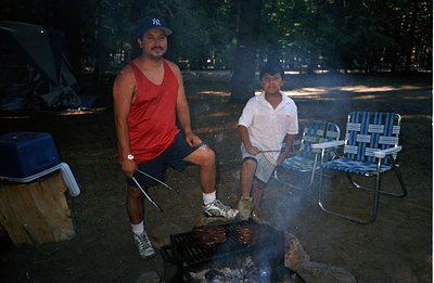 A man and boy grill food over a campfire. The adult wears a red tank top and baseball cap, while the boy is in a short-sleeve...
