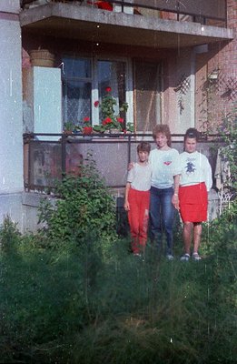 Three children stand in overgrown grass before a block of flats. The building features a balcony adorned with potted geranium...