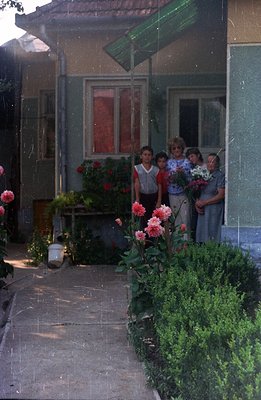 A group of four young people stands in a doorway, holding bouquets of flowers. The scene appears to be a residential courtyar...