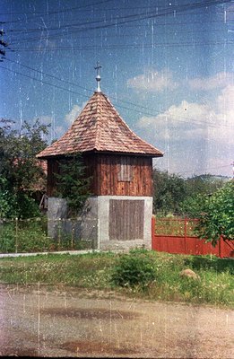Ornate, timber-framed bell tower with a pyramidal roof topped by a cross, sits upon a concrete base. Traditional Romanian arc...