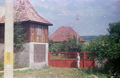 A weathered, tiled-roof building stands adjacent to a red-painted picket fence and a figure stands beyond. The architecture s...