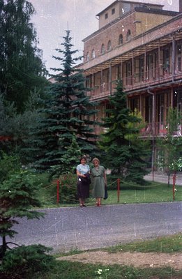 A 1970s color photograph depicts two women in knee-length dresses walking along a paved path alongside a grand, Soviet-era sa...