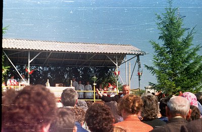 Outdoor religious ceremony captured from a crowd perspective. A robed clergy stands at an altar beneath a simple, corrugated ...