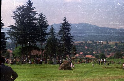 A hillside meadow hosts a gathering, likely a festival or public event. Traditional dress is visible among the attendees. Bac...