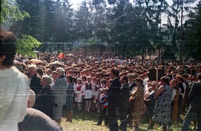 Large outdoor gathering with crowds of people, some in formal attire. Visible are children wearing white shirts and red neckt...
