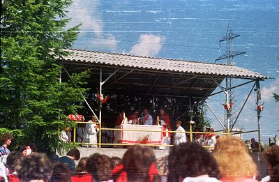 Outdoor religious ceremony takes place under a simple, corrugated metal shelter. Several figures in white robes gather around...