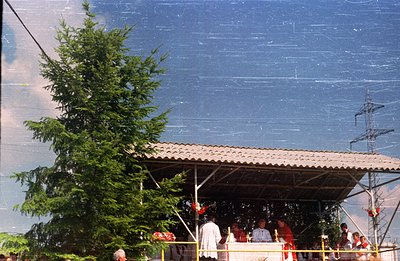 A covered platform with a corrugated metal roof hosts a gathering of people, possibly a religious ceremony or public event. A...