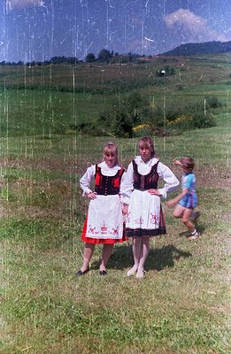 Two young women in traditional folk dress stand facing the camera amidst a grassy hillside. Detailed black & red embroidered ...