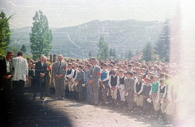 A large group of formally dressed boys stand in a line outdoors, likely schoolchildren. Men in suits observe the group, some ...