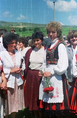 Three women in traditional folk attire stand in a field alongside others at an outdoor gathering. The woman in the center wea...