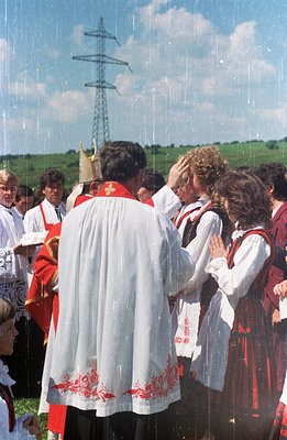 A ceremonial blessing in a rural setting, likely a folk festival. A priest in embroidered vestments blesses a young couple in...