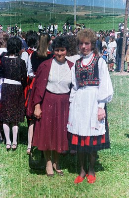 Two women stand in a field of green grass, both wearing traditional folk dress. The woman on the left wears a red skirt and j...