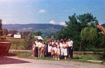 Group portrait of a large family posed outdoors before a backdrop of rolling hills and traditional rural buildings. Subjects ...