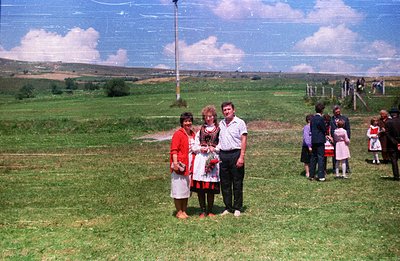 A group portrait on a grassy hillside. Three adults stand prominently in front of a wider gathering, likely a community event...