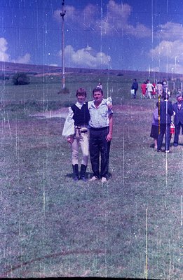 A man and woman in casual attire stand close together outdoors. The woman wears riding breeches and a ruffled blouse, suggest...