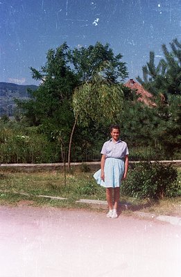 A young woman stands on a gravel path beside a willow tree, likely in Bulgaria. She wears a light blue, patterned dress and w...