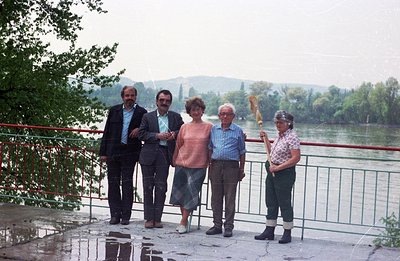 Four individuals pose near a riverbank, likely a holiday or family snapshot. Man with beard (left) in a suit, another in a bl...