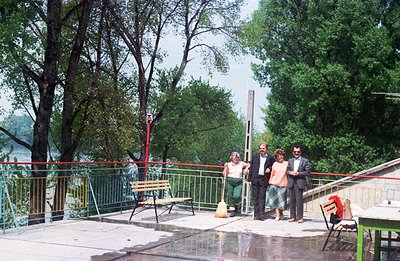 Outdoor scene: Four people stand on a patio overlooking a river, likely a resort or public space. Architecture includes a low...
