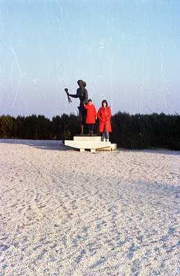 A bronze statue depicting a man in historical military attire stands on a stone pedestal. Two children in matching red parkas...