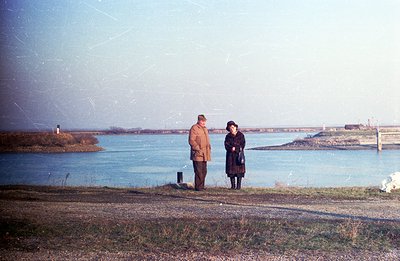 A color photograph depicts a couple standing on a grassy verge overlooking a wide body of water, possibly a bay or inlet. The...