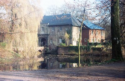 A historic mill building, constructed of brick and stone, stands beside a placid, reflective pond. The structure features mul...