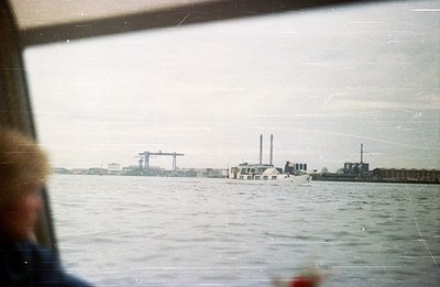 An industrial harbor scene: a boat traverses a body of water, framed by factory structures & cranes along the distant coastli...