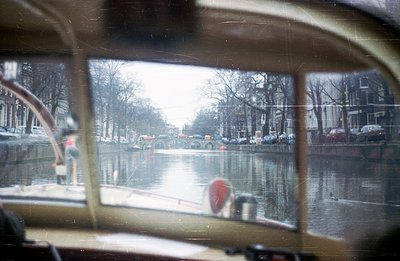 View from within a vehicle, likely a boat, showing a canal scene. Buildings line the waterway, reflecting in the water. Trees...