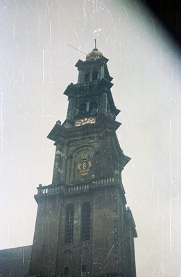 A tall, weathered stone church tower dominates the frame, featuring a clock face and ornate detailing. Visible signs of aging...