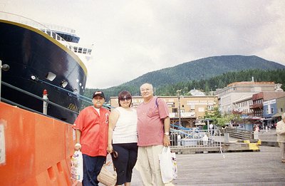 A family stands on a pier, posed in front of a large cruise ship and a backdrop of forested mountains. Appears to be a touris...