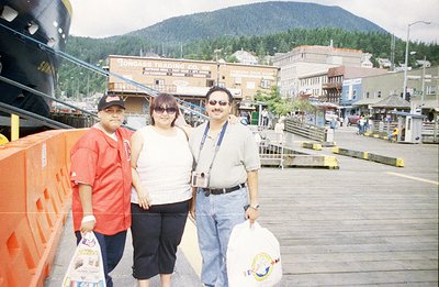 A family of three poses near a pier in an Alaskan coastal town. Visible signs identify "Tongass Trading Co." and "Icy Strait ...