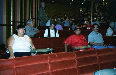 Interior view of a cinema with patrons seated in tiered red velvet seating. A group of five people are prominently featured i...