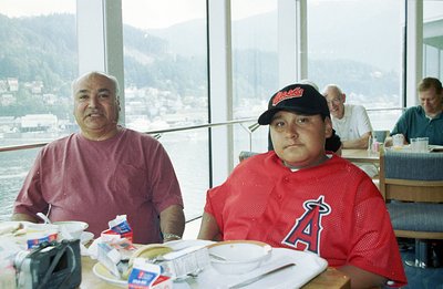 Two men seated at a table by large windows overlooking a coastal landscape with mountains in the distance. The man on the lef...