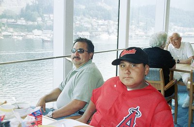 Two men seated at a restaurant with large windows overlooking a harbor and mountainous coastline. One wears sunglasses and a ...