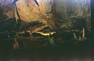 Interior of a cave, showcasing rock formations and illuminated by artificial lighting. A dark path leads deeper into the cave...