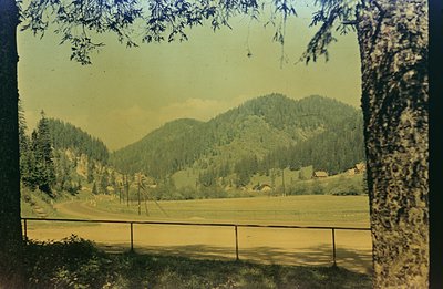 A wide, expansive view across a grassy field toward forested mountains. A section of dark wooden fencing runs along the foreg...