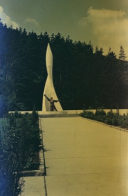 Monumental sculpture of a figure with outstretched arms atop a stark, modernist tower. Surrounded by lush evergreen foliage, ...